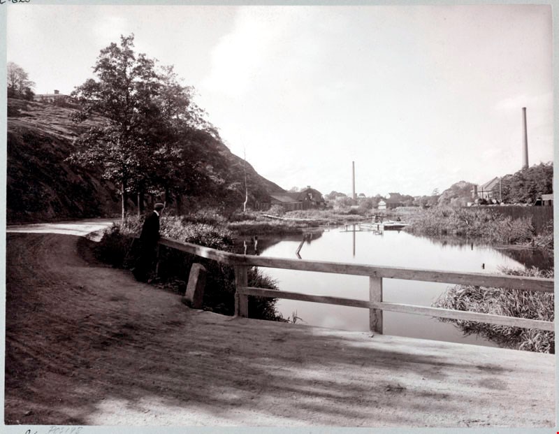 Hammarby sjös utlopp vid Danviken 1915 innan kanalen började grävas ut. Sjön var delvis igenväxt. Till höges syns Liljeholmens fabriker på Södermalmssidan. Vägen slingrar sig vidare runt Danviksklippan mot Värmdö till vänster i bilden. Fotograf Aktiebolaget Saltsjöqvarn. Nacka lokalhistoriska arkiv.