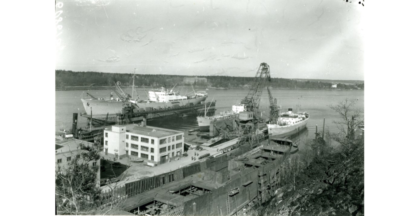 De båda husen på bilden från 1949 står kvar och stapelbädden syns tydligt i bildens nederkant. Det ljusa huset var snickeri- och timmermansverkstad. Huset till vänster var verkstadskontoret. Fotograf Stig Herbst. Sjöhistoriska Fo 65448C.