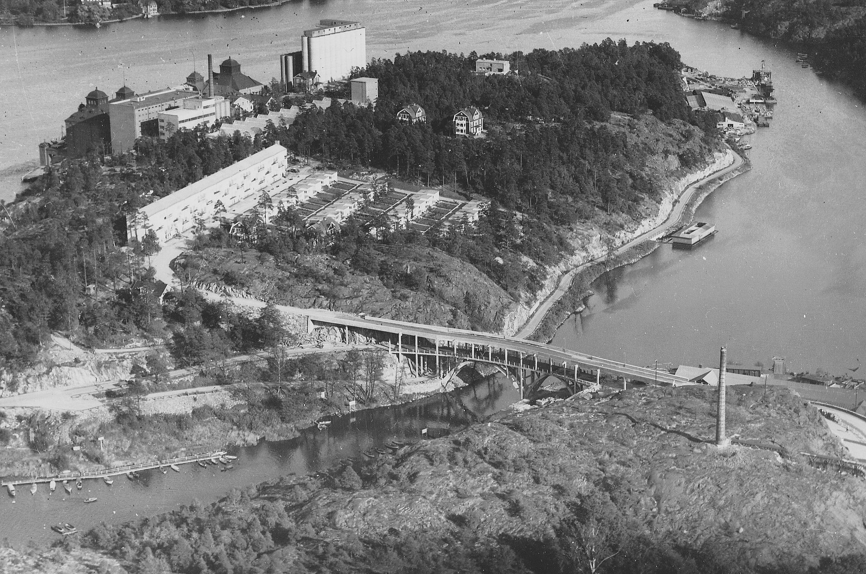 Kvarnholmen 1932 med bron och Hästholmssundet, innan det fylldes igen. Det så kallade smalhuset är byggt, liksom radhusen nedanför. EPA-huset är ännu inte byggt. Småbåtsbryggorna syns tydligt i sundet. Nere till höger står skorstenen som släppte ut gaser från superfosfatfabriken. Där byggdes senare Finnboda varvs personalbostäder. Fotograf Oscar Bladh.